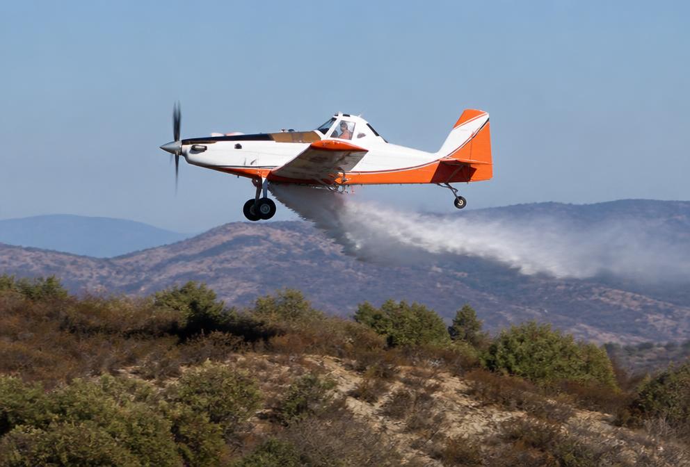 Avión monomotor de extinción, blanco y naranja, soltando una línea estrecha de agua sobre matorral seco mediterráneo.
