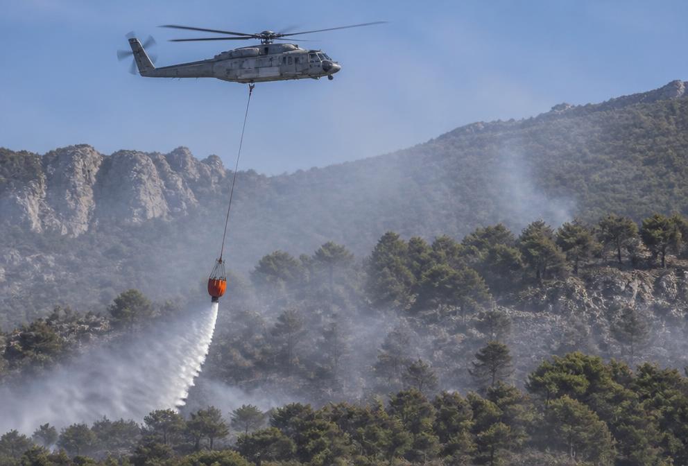 Helicóptero de extinción con helibalde suspendido descargando agua sobre una ladera arbolada con humo.